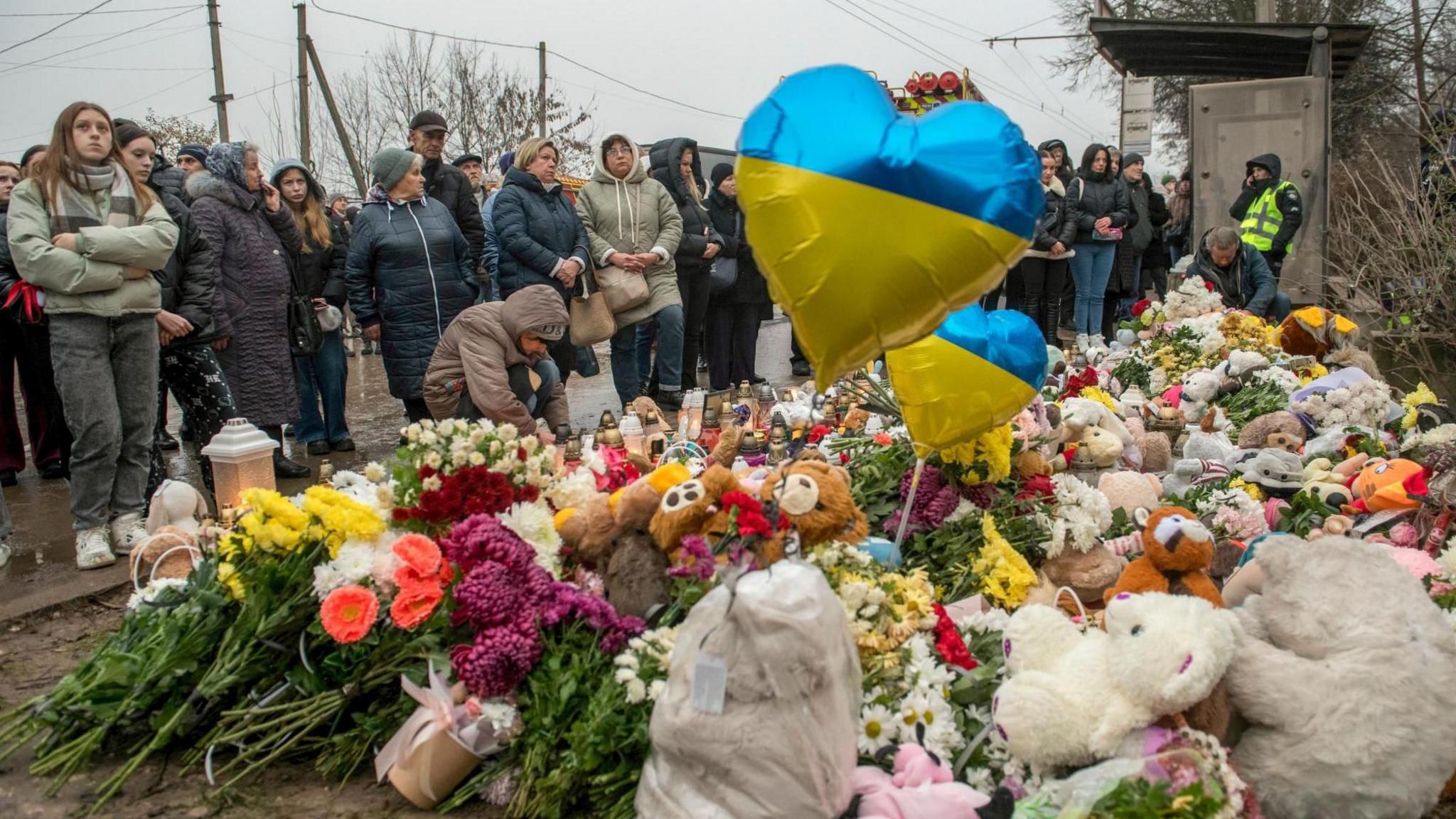 Dhjetëra njerëz, të veshur me kapele dhe xhaketa të mëdha, mblidhen rreth një shtrati me lule të vendosura në tokë si një memorial për viktimat e një sulmi rus. Midis luleve, ka lodra përqafuese dhe tullumbace me flamurin ukrainas në formën e një zemre.