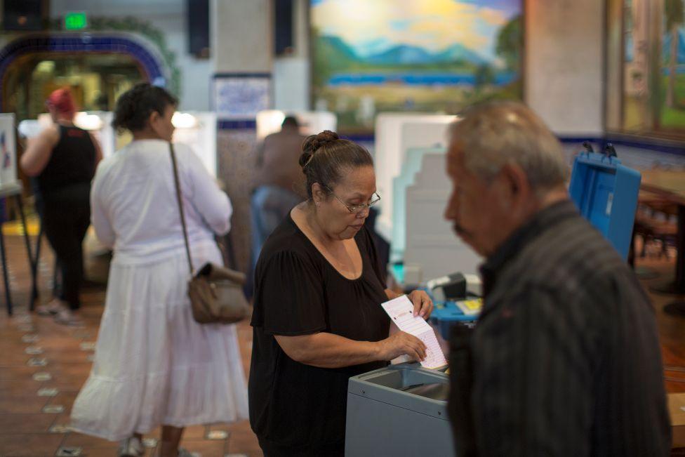 Grua që mban fletë votimi pranë qendrës së votimit me një burrë tjetër në plan të parë