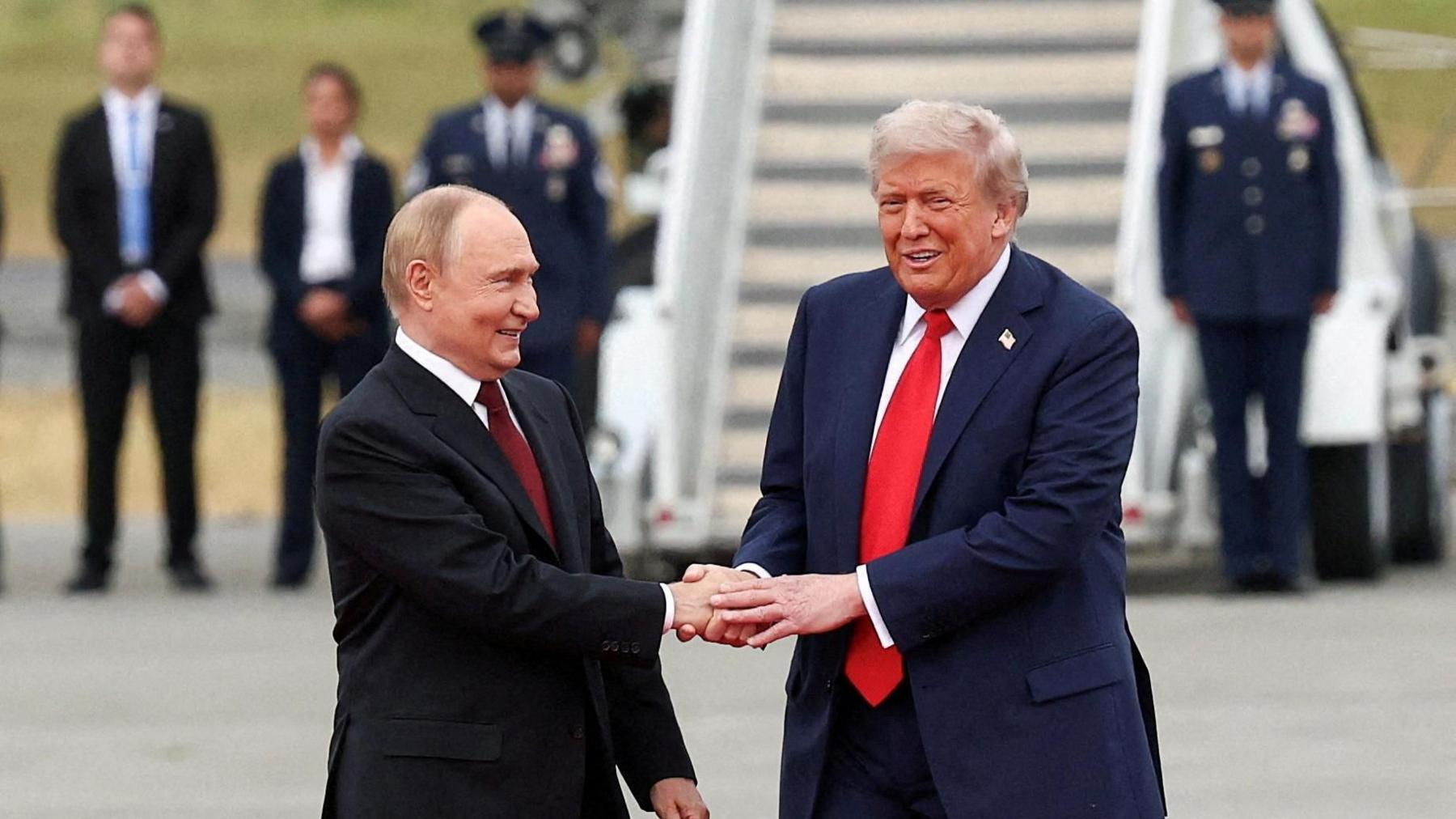 Trump dhe Putin qëndrojnë në pistën e aeroportit në Alaskë përpara samitit të tyre, duke shtrënguar duart dhe duke buzëqeshur. Pas tyre qëndron një rresht burrash dhe grash të veshur me uniformë, dhe një shkallë e madhe që të çon drejt një avioni. Putin ka veshur një kostum të zi, këmishë të bardhë dhe kravatë të kuqe të errët, dhe Trump ka veshur një kostum blu të errët, këmishë të bardhë dhe kravatë të kuqe.