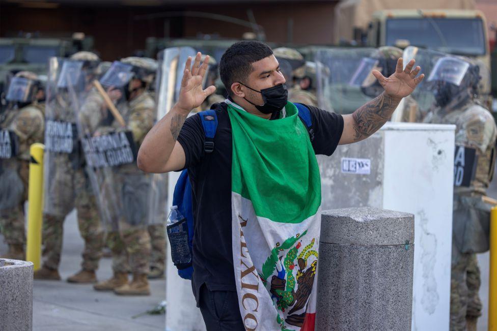 Një burrë i mbështjellë me flamurin e Meksikës me krahët e ngritur ecën përpara ushtarëve të Gardës Kombëtare të Kalifornisë me mburoja, ndërsa protestat vazhdojnë në një zonë prej afërsisht një milje katrore në qendër të Los Angeles në qershor 2025.