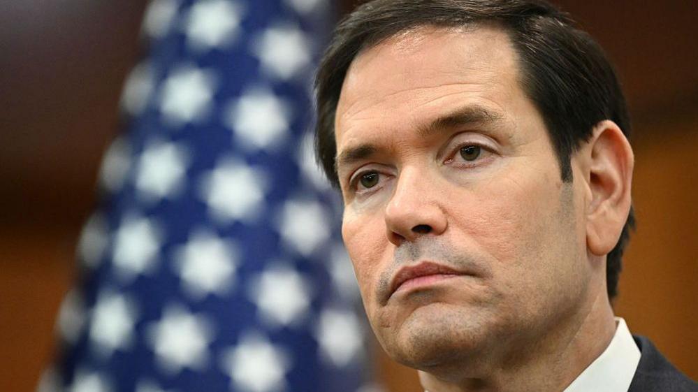 Një imazh i Sekretarit të Shtetit të SHBA-së, Marco Rubio, duke folur në mikrofon i veshur me një kostum blu të errët me kravatë të kuqe.