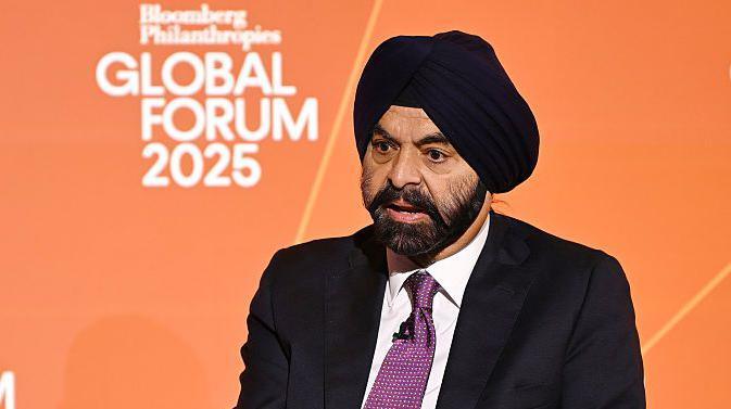 Një imazh i Ajay Bangës duke folur para një sfondi portokalli, i veshur me një çallmë të zezë dhe kostum të zi me një kravatë vjollcë.