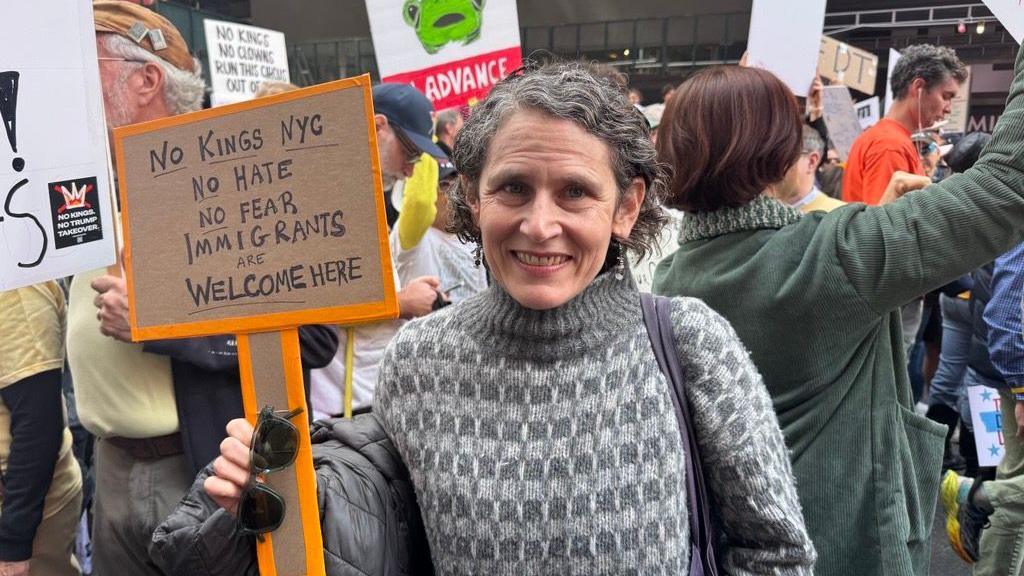 Beth Zasloff duke buzëqeshur para kameras para turmës së protestuesve, duke mbajtur një pankartë që thotë "Pa mbretër në Nju Jork, pa urrejtje, pa frikë, emigrantët janë të mirëpritur këtu".