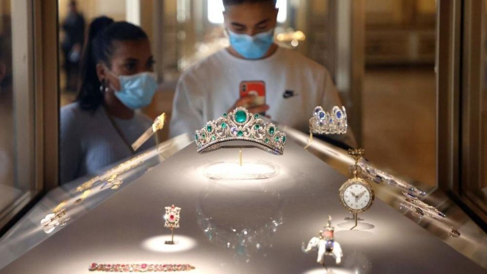 Vizitorët që mbajnë maska ​​në fytyrë shikojnë Diademën e Dukeshës së Angouleme të ekspozuar në një dritare të shfaqur në galerinë Apollo në Muzeun e Luvrit.