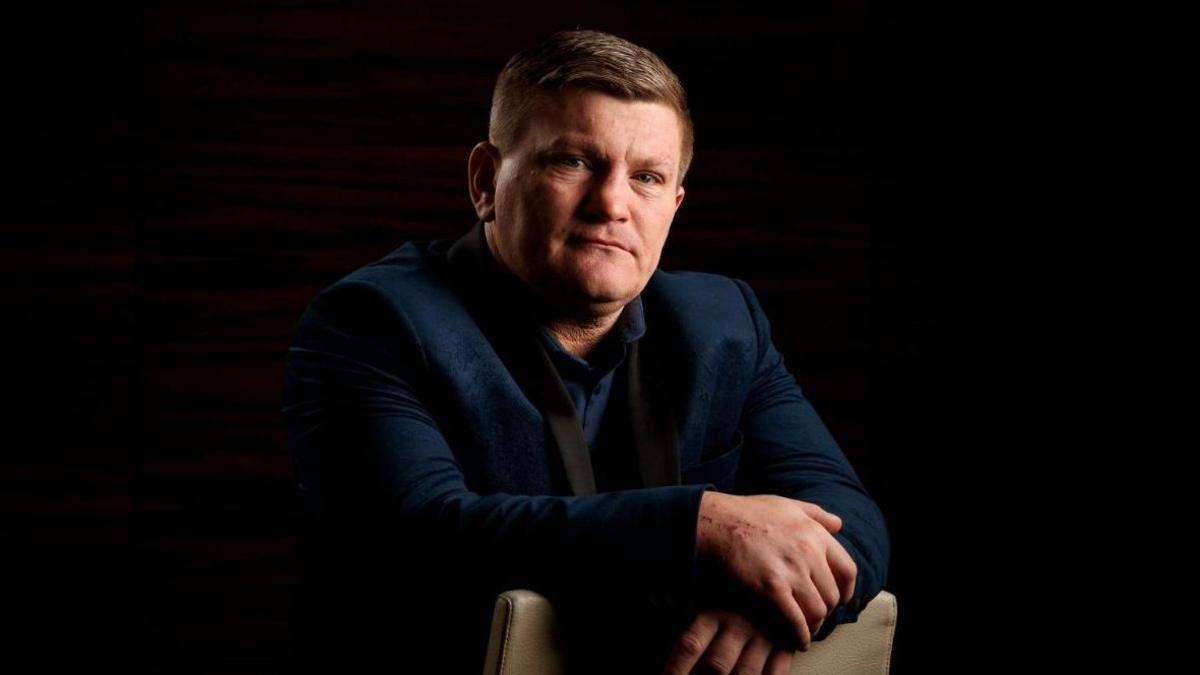 Ricky Hatton po e shikon drejt e në kamerë në këtë fotografi të pozuar. Ai ka veshur një kostum dhe një këmishë ngjyrë blu të errët.