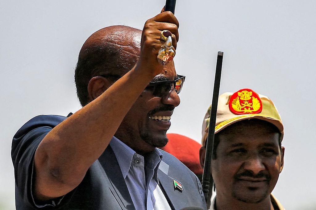Omar al-Bashir (majtas) tund një bastun ndërsa mban një fjalim në selinë e paraushtarakëve të Forcave të Mbështetjes së Shpejtë (RSF) në Umm al-Qura në shtetin e Darfurit Jugor, ndërsa komandanti i RSF-së, Mohamed Hamdan Daglo, i veshur me një kapele, shikon.