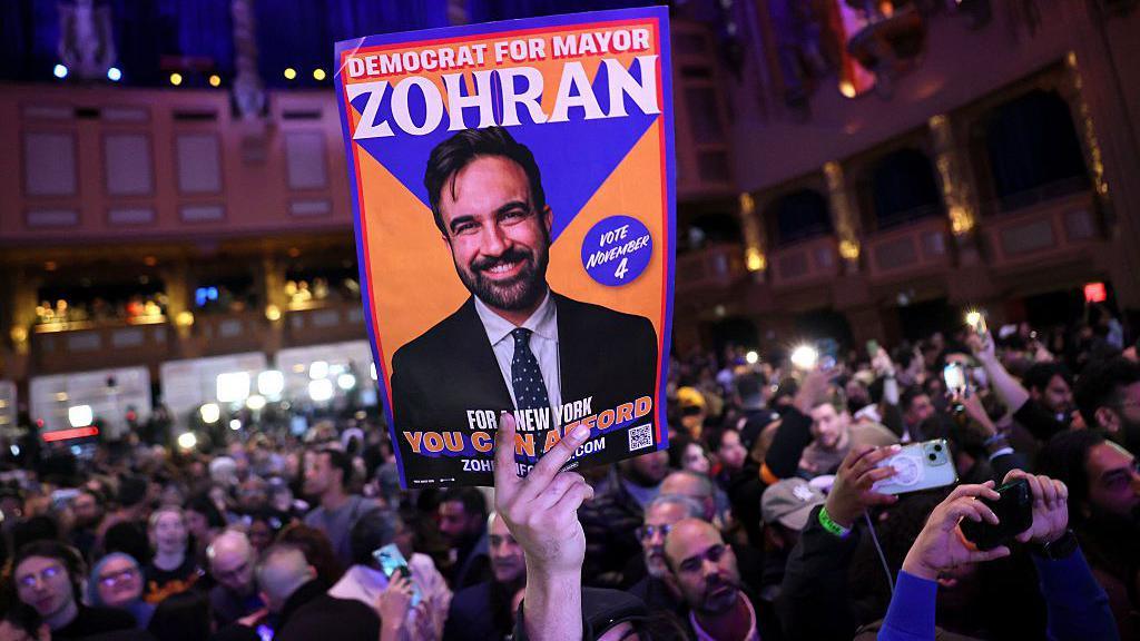 Dora e një personi të bardhë që mban një tabelë portokalli me foton e Mamdanit mbi të, ku shkruhet "Demokrat për Kryetarin e Bashkisë Zohran" mbi një det me kokash dhe kamerash në një sallë të ndriçuar dobët.