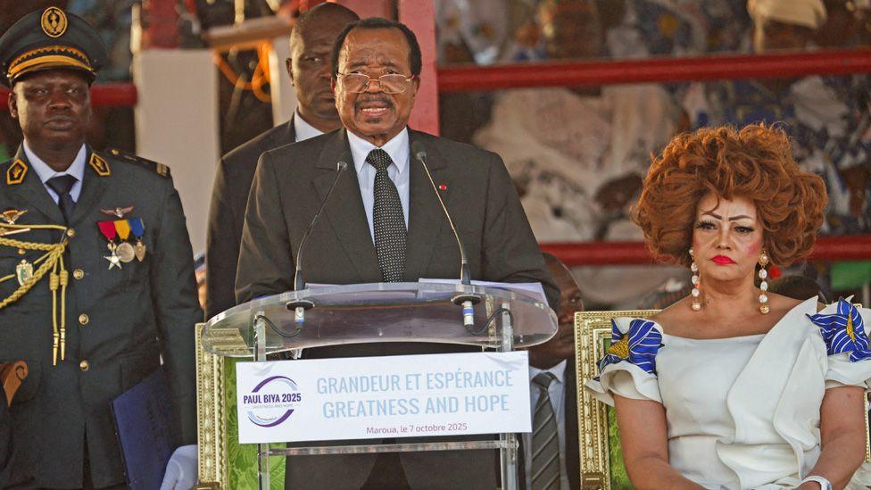 Presidenti Paul Biya flet në një foltore i veshur me kostum. Gruaja e tij Chantal ulet pranë tij me një fustan të bardhë me zbukurime me lule në shpatulla. Pas tyre qëndron një oficer ushtarak, një zyrtar tjetër dhe pas tyre mbështetës të presidentit të Kamerunit - Maroua, Kamerun, 7 tetor 2025.