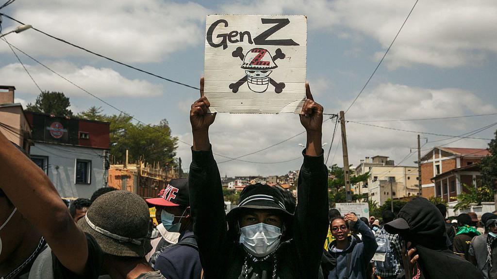 Një protestues me fytyrën e mbuluar nga një maskë mban lart një poster që thotë 'Gjenerata Z'
