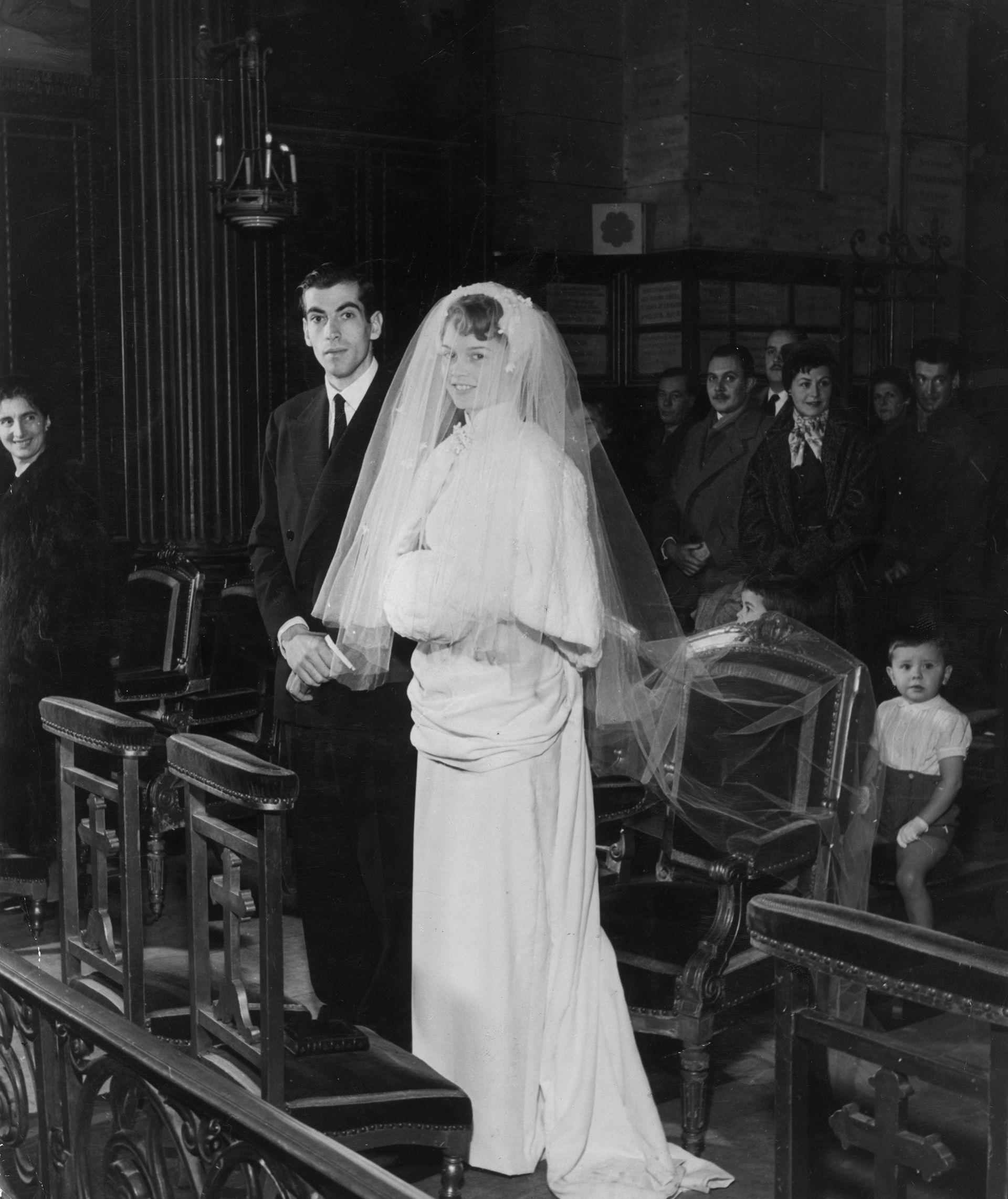 Brigitte Bardot dhe Roger Vadim, duke qëndruar te altari gjatë ceremonisë së tyre të dasmës në dhjetor 1952. Fytyra e Bardot është e mbuluar nga një vello e gjatë dhe Vadim mban veshur një kostum dhe kravatë të errët.