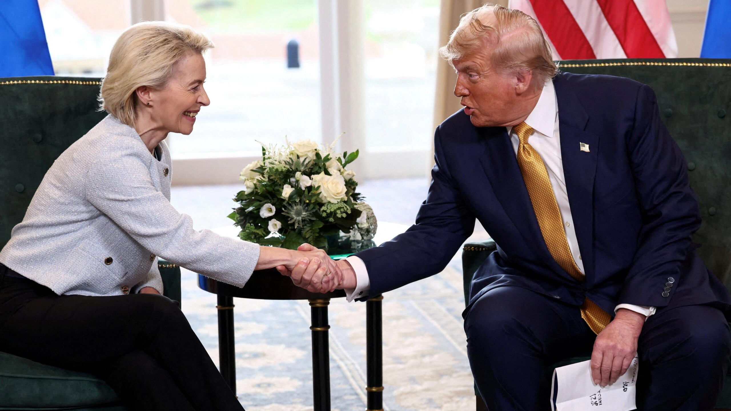 Von der Leyen, me një xhaketë të bardhë të shkurtër dhe pantallona të zeza, e shtrin dorën përpara një tavoline anësore me një aranzhim lulesh të bardha për të kapur dorën e Trump, i cili është me kostum blu dhe kravatë të artë dhe mban letra në dorën tjetër.