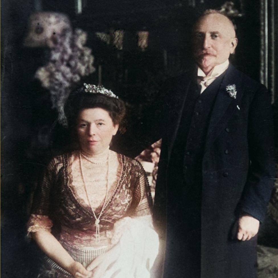 Fotografi historike e ngjyrosur e Olgës dhe Heinrich Eisnerit me veshje zyrtare. Olga është ulur, e veshur me një fustan dantelle me një diademë dhe duke mbajtur një copë të bardhë. Heinrich qëndron pranë saj i veshur me një kostum të errët me një butonniere. Sfondi përfshin aranzhime me lule dhe dekor të zbukuruar, që tregojnë një ambient elegant.