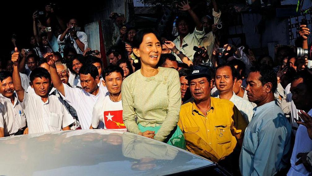 Aung San Suu Kyi (C) buzëqesh ndërsa mbërrin në selinë e Lidhjes Kombëtare për Demokraci (NLD) në Yangon më 15 nëntor 2010. Ajo është e rrethuar nga një turmë.
