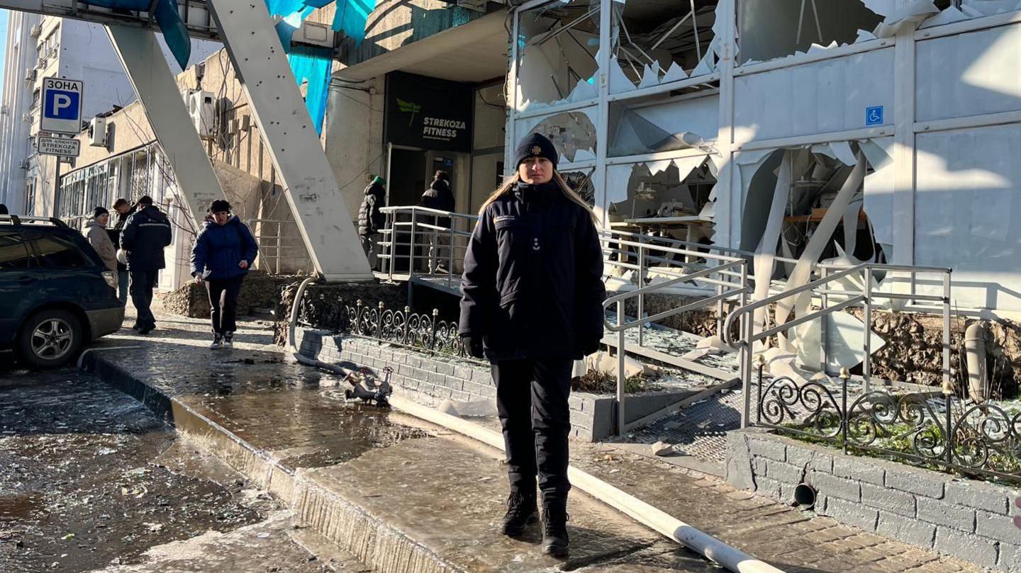 Një grua e veshur me xhaketë blu dhe kapelë qëndron përpara një ndërtese të dëmtuar