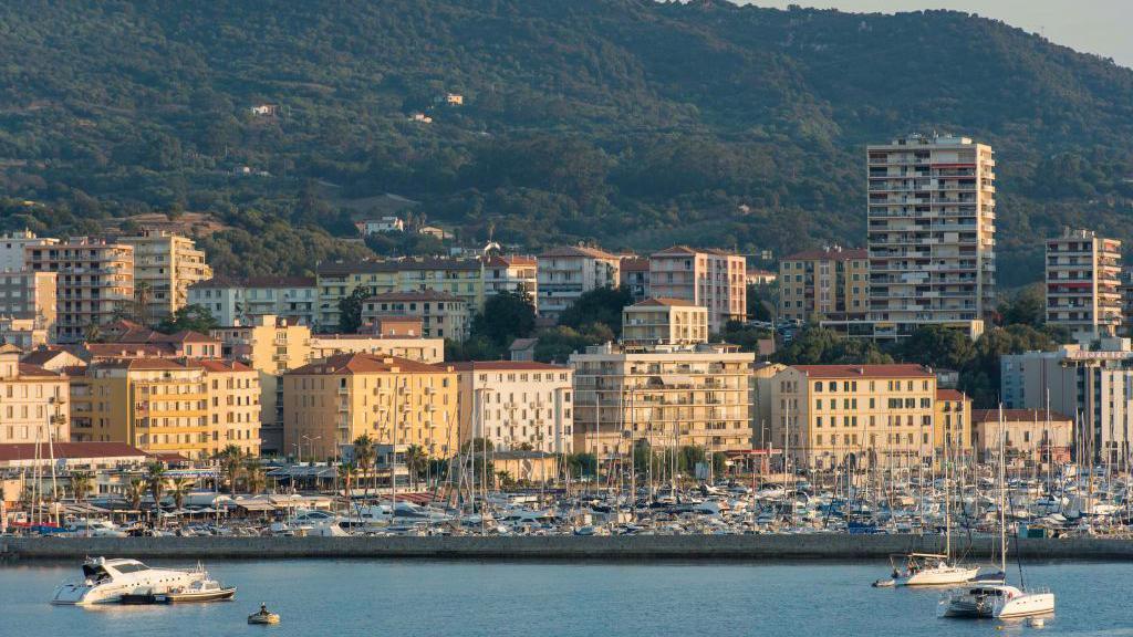 Dielli i mbrëmjes shkëlqen mbi blloqet e verdha të apartamenteve në bregdetin e Ajaccio-s, me marinën dhe anijet përpara