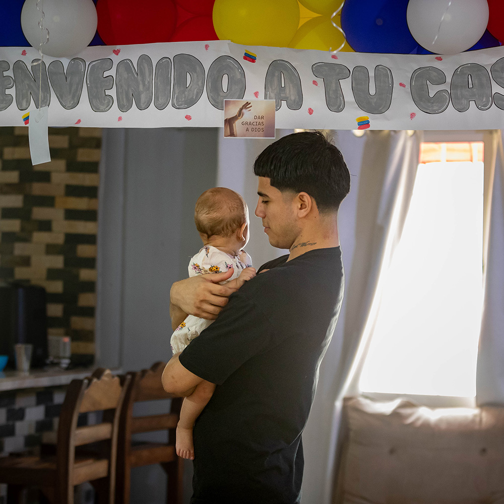 Joén Suarez, i veshur me një bluzë të zezë, mban vajzën e tij të vogël në shtëpinë e tij në Venezuelë. Në sfond ka tullumbace të kuqe, të bardha, blu dhe të verdha dhe një tabelë që thotë "Mirë se vini në shtëpi" në spanjisht.