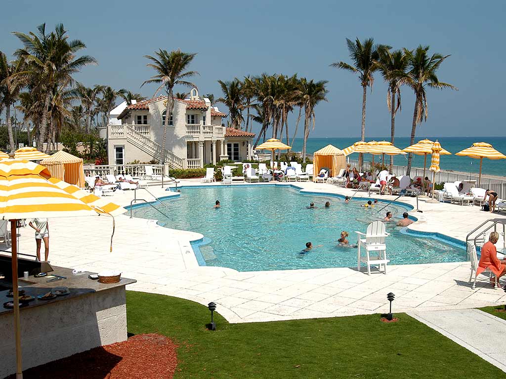 Zona e pishinës së jashtme në Mar-a-Lago's Beach Club me pamje nga oqeani. Pamja paraqet një pishinë të madhe, me formë të çrregullt, të rrethuar nga shezlongje dhe tavolina të bardha, të hijezuara nga çadra me vija të verdha dhe të bardha. Palmat shtrihen përgjatë skajit të pronës, dhe oqeani bruz është i dukshëm në sfond. Një ndërtesë në stil mesdhetar me mure të bardha prej llaçi dhe çati me pllaka të kuqe qëndron pranë pishinës.