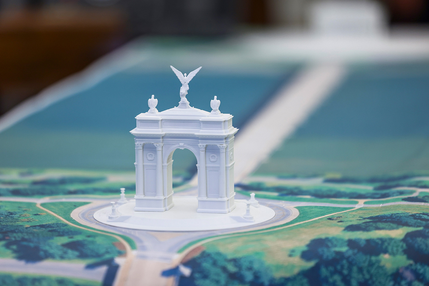 Pamje nga afër e një modeli arkitektonik të bardhë të 'Arc de Trump', të vendosur në një hartë të terreneve të peizazhuara. Dizajni përfshin një hark qendror të rrethuar nga dy harqe më të vogla, detaje të zbukuruara dhe një figurë me krahë sipër strukturës. Modeli qëndron mbi një bazë rrethore me shtigje që shtrihen nga jashtë.