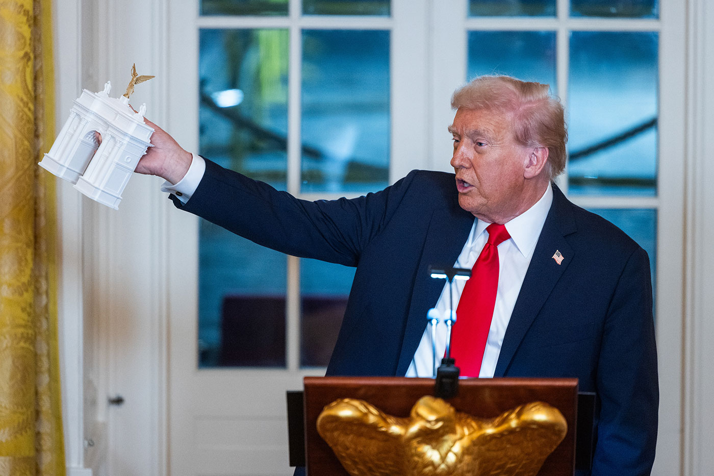 Donald Trump i veshur me kostum të errët dhe kravatë të kuqe, duke qëndruar në një podium me një emblemë shqiponje të artë, duke mbajtur lart një model të bardhë në shkallë të harkut neoklasik.