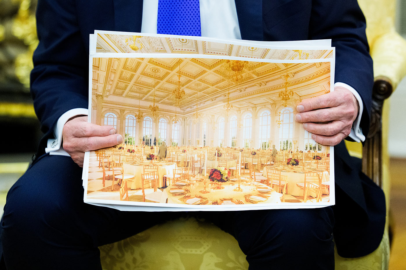 Foto e Donald Trump duke mbajtur një printim të madh të brendshëm të sallës së vallëzimit. Dizajni përmban dritare të larta me hark, tavane me arka të zbukuruara me zbukurime ari, llambadarë dhe tavolina të rrumbullakëta të vendosura me karrige dhe dekorime qendrore me lule.